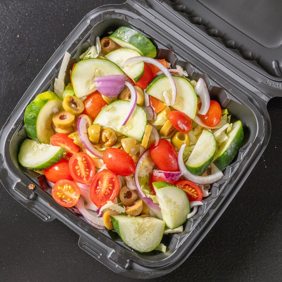Fresh garden salad with lettuce, cucumber, tomatoes, red onion, and sliced olives in a black takeout container.