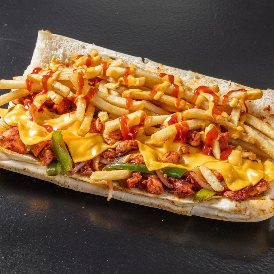 Loaded sandwich topped with fries, grilled meat, peppers, onions, melted cheese, ketchup, and creamy sauce on a dark surface.
