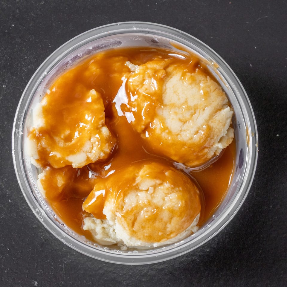 Three dumplings covered in glossy brown gravy sit in a clear plastic container on a dark countertop.