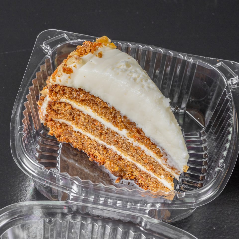 Slice of layered carrot cake with white frosting and crumb topping in a clear plastic container.