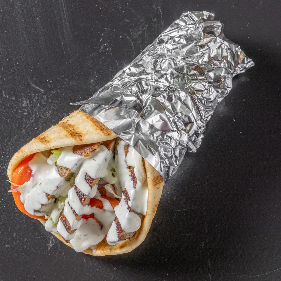 Gyro-style pita wrap with grilled meat, lettuce, tomatoes, onions, and creamy white sauce, partially wrapped in foil.