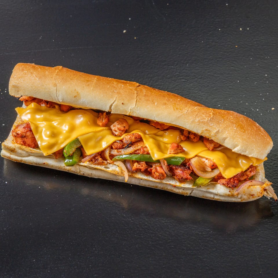 Long toasted sandwich filled with grilled meat, melted cheddar cheese, green peppers, onions, and bacon on a black surface.