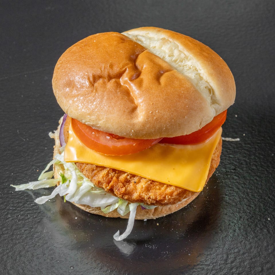 Crispy chicken burger with melted cheese, lettuce, tomato, and onion on a soft bun against a dark background.