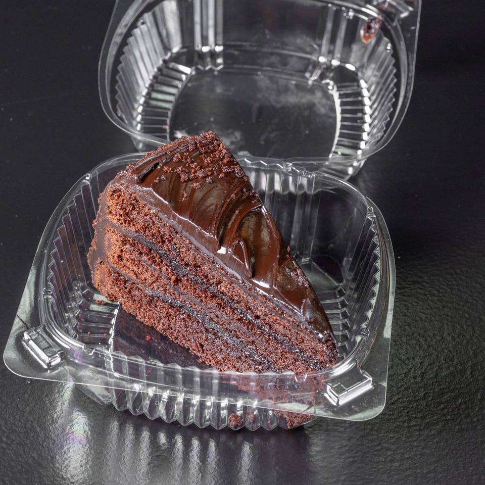 Slice of layered chocolate cake with glossy frosting and chocolate crumbs in an open plastic container.