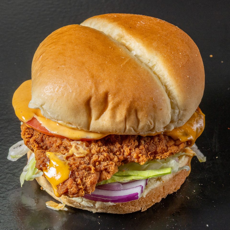 Close-up of a crispy fried chicken burger with melted cheese, lettuce, tomato, onion, and sauce on a toasted bun.