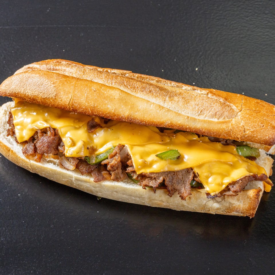 Toasted baguette sandwich filled with shaved steak, melted cheese, peppers, and onions on a dark surface.
