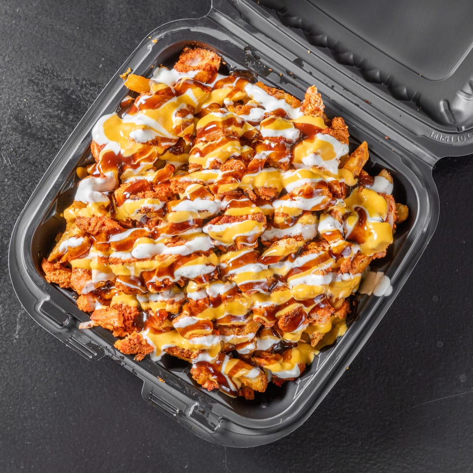 Loaded fries topped with crispy chicken, melted cheese, white sauce, and barbecue sauce in a black takeout container.