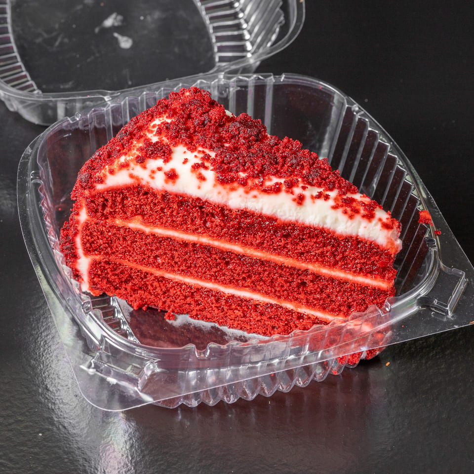 Slice of red velvet cake with creamy white frosting layers and red crumbs in a clear plastic container.