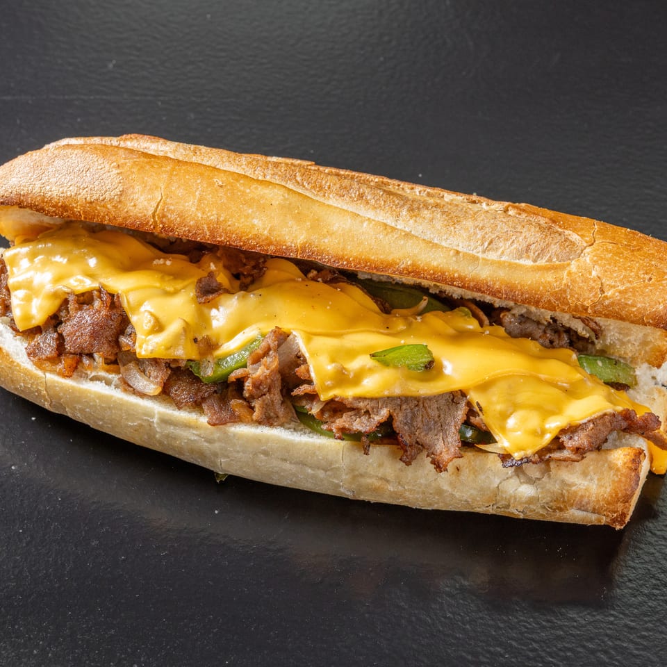 Philly cheesesteak sandwich with sliced beef, melted yellow cheese, peppers, and onions on a crusty baguette.